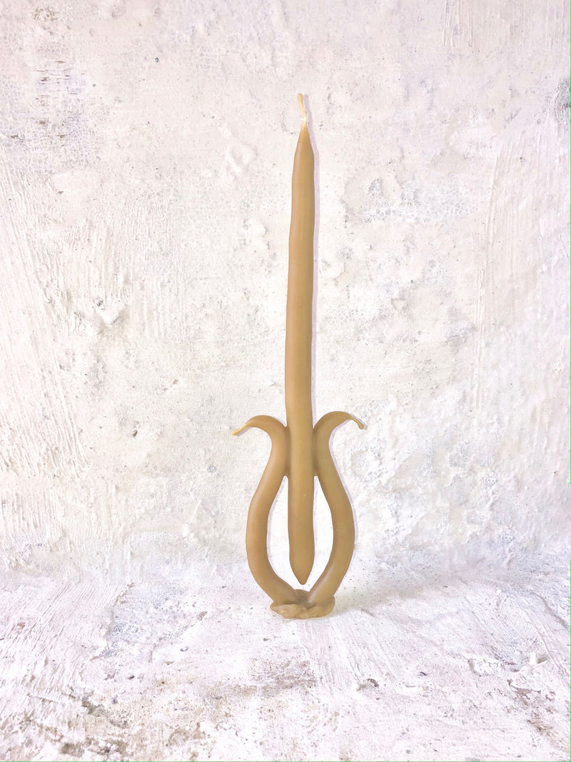A unique beige hand-dipped beeswax Candle 036 by Ola Lewczyk with an artistic, double-curving base stands against a textured white background. The candle's elegant and fluid shape suggests movement, resembling a treble clef.
