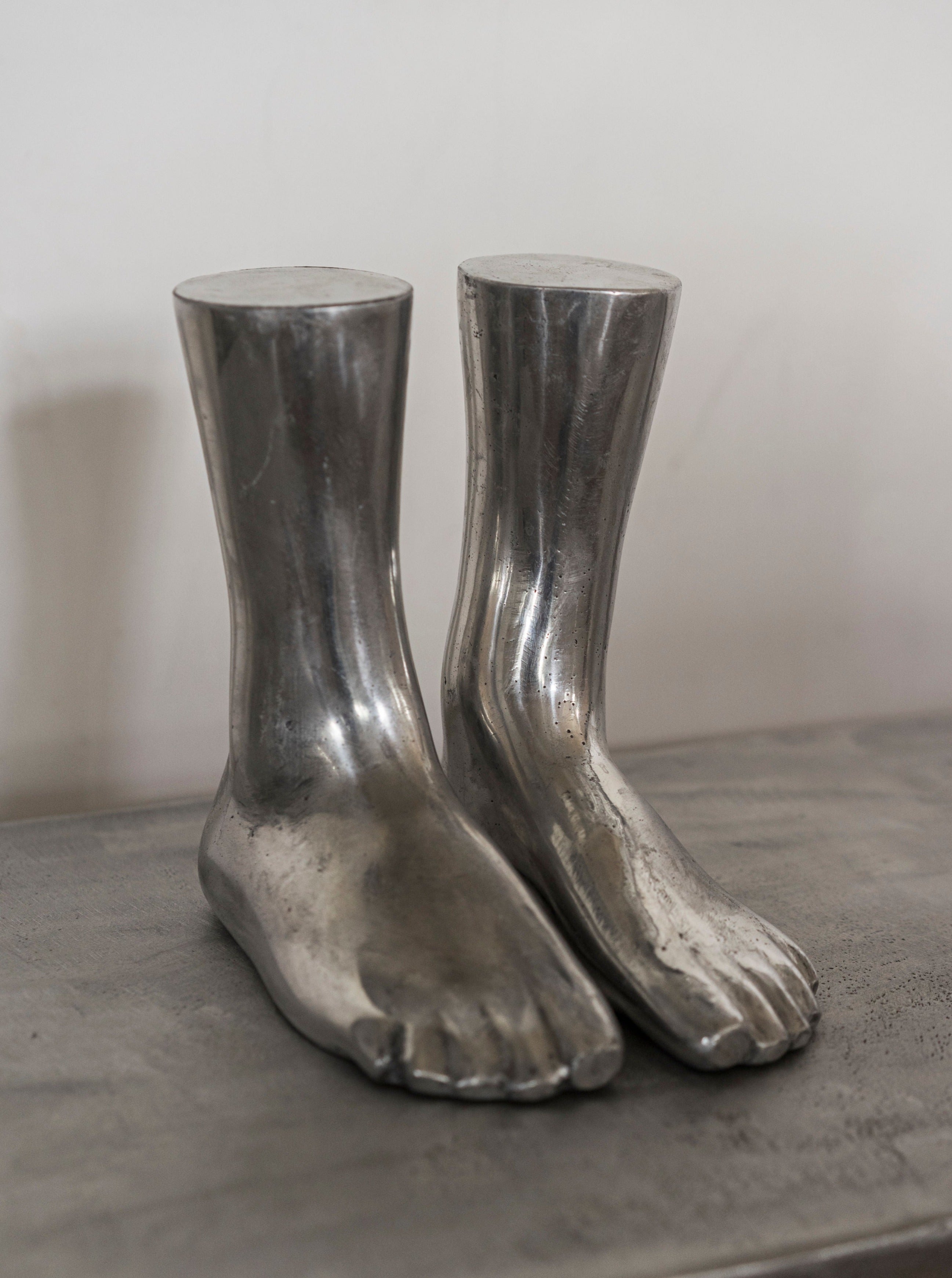 A pair of Rooms Studio casted aluminum feet sculpture boots designed to resemble human feet, with detailed toes and shin-high length, displayed on a grey surface against a neutral background.