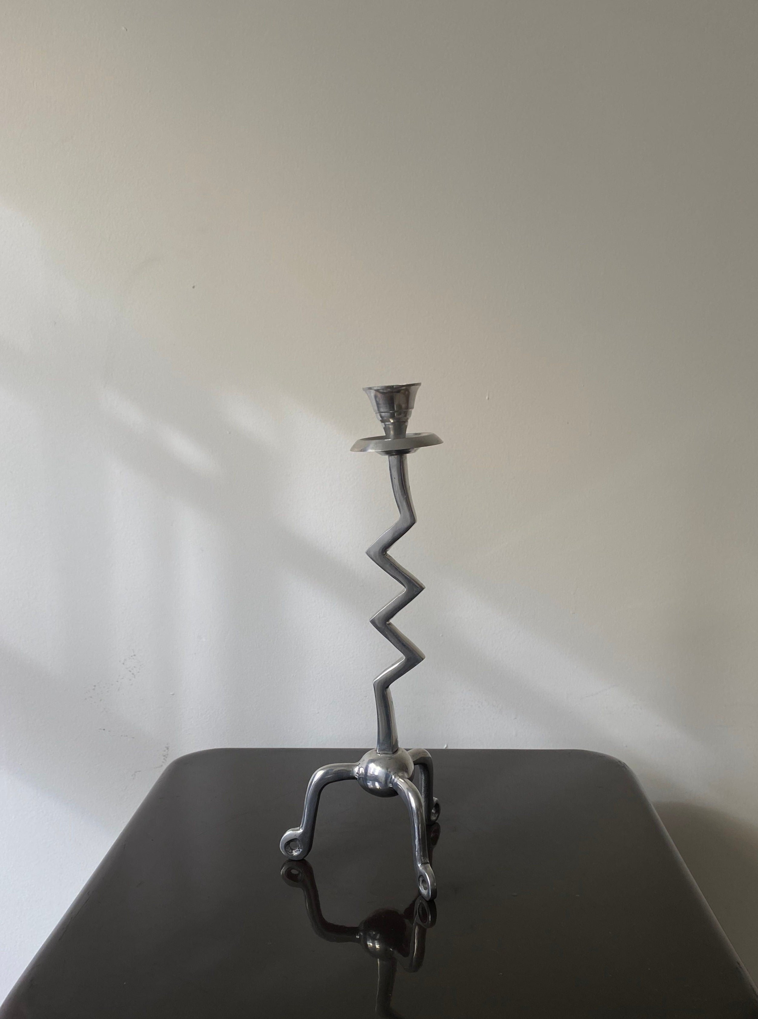 A Vintage Zigzag Candle Holder by Les Objets Amsterdam with a unique zigzag stem and four-legged base, placed on a dark surface against a light grey wall with gentle shadow patterns.