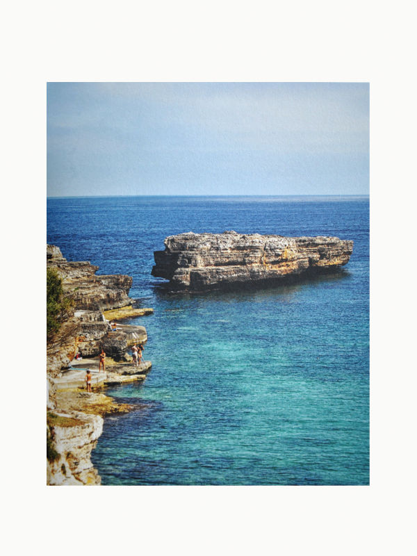 A serene coastal scene featuring clear turquoise water and layered rock formations, as depicted in *The Orange Set of 3 Interior Design and Art Books* by Maison Plage, evokes creative inspiration. People are seen standing and observing the ocean from the rocky shoreline. The clear blue sky above enhances the tranquil atmosphere captured in these beautifully crafted books.