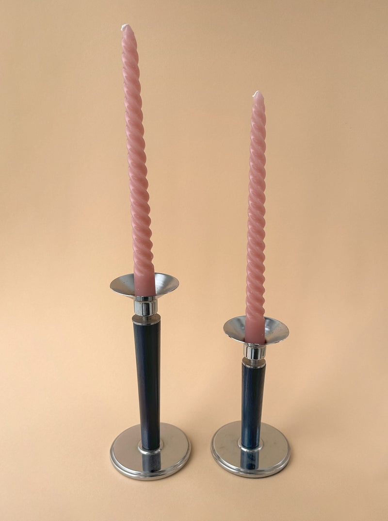 Two pink spiral candles in vintage silver Dodo Vintage candleholders with black handles, arranged symmetrically on a light peach background.