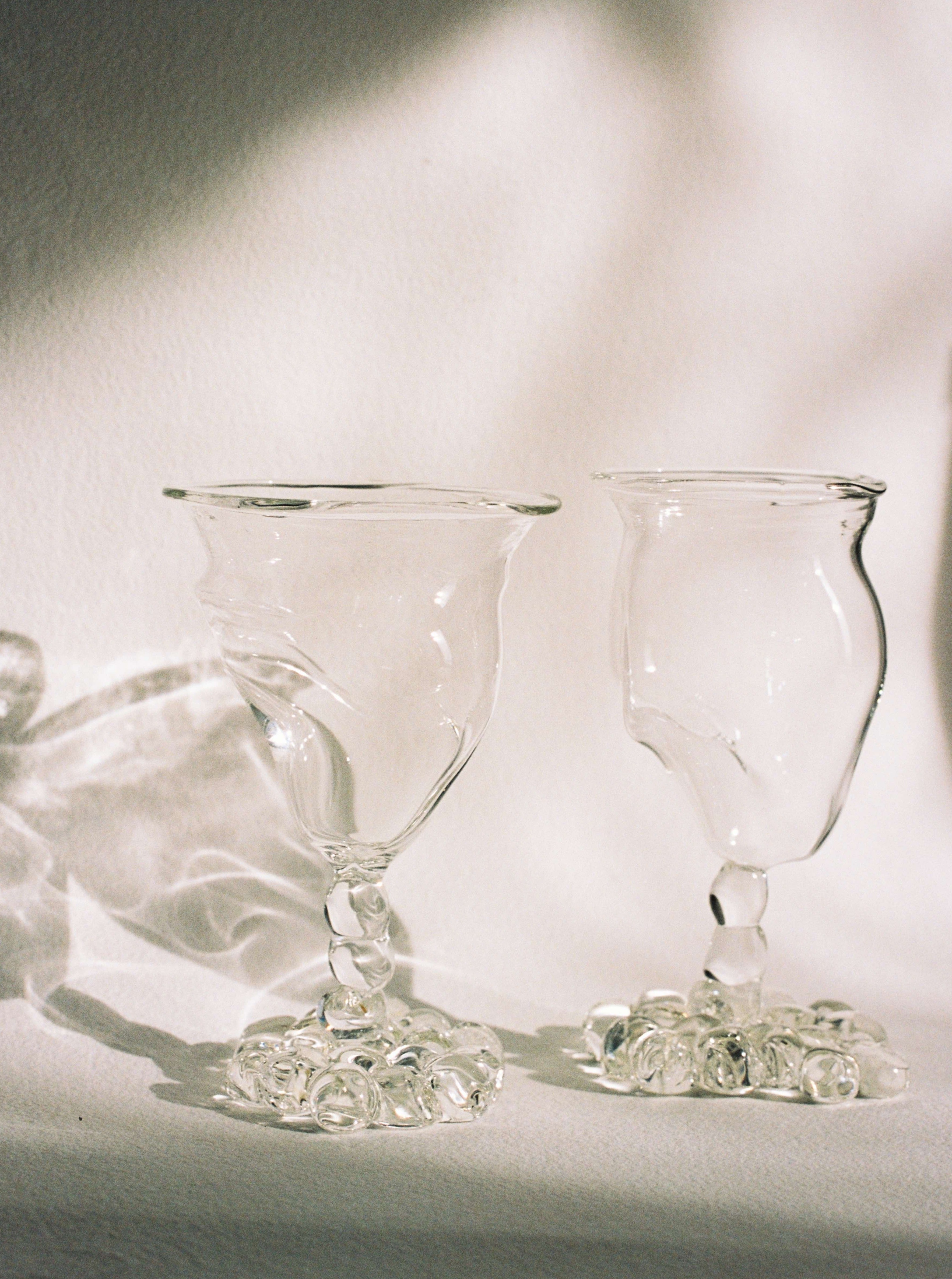 Two Justine Menard Mouthblown Wine Glasses on a table, illuminated by soft light casting shadows, surrounded by scattered clear pebbles, giving a serene and elegant look.