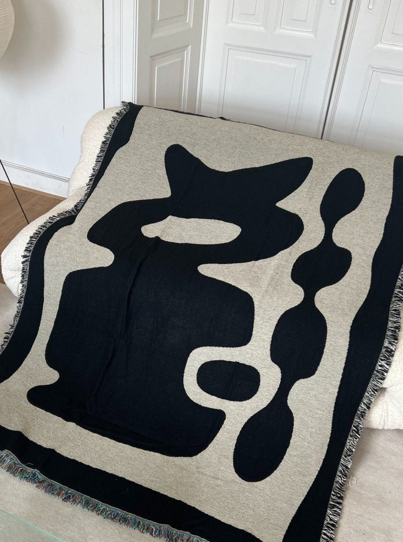 A modern Kozu Jacquard Throw from 2222Studio, with an abstract design in black and gray, laid out on a wooden floor against a white door background. The throw features bold, irregular shapes and serves as a decorative accent.