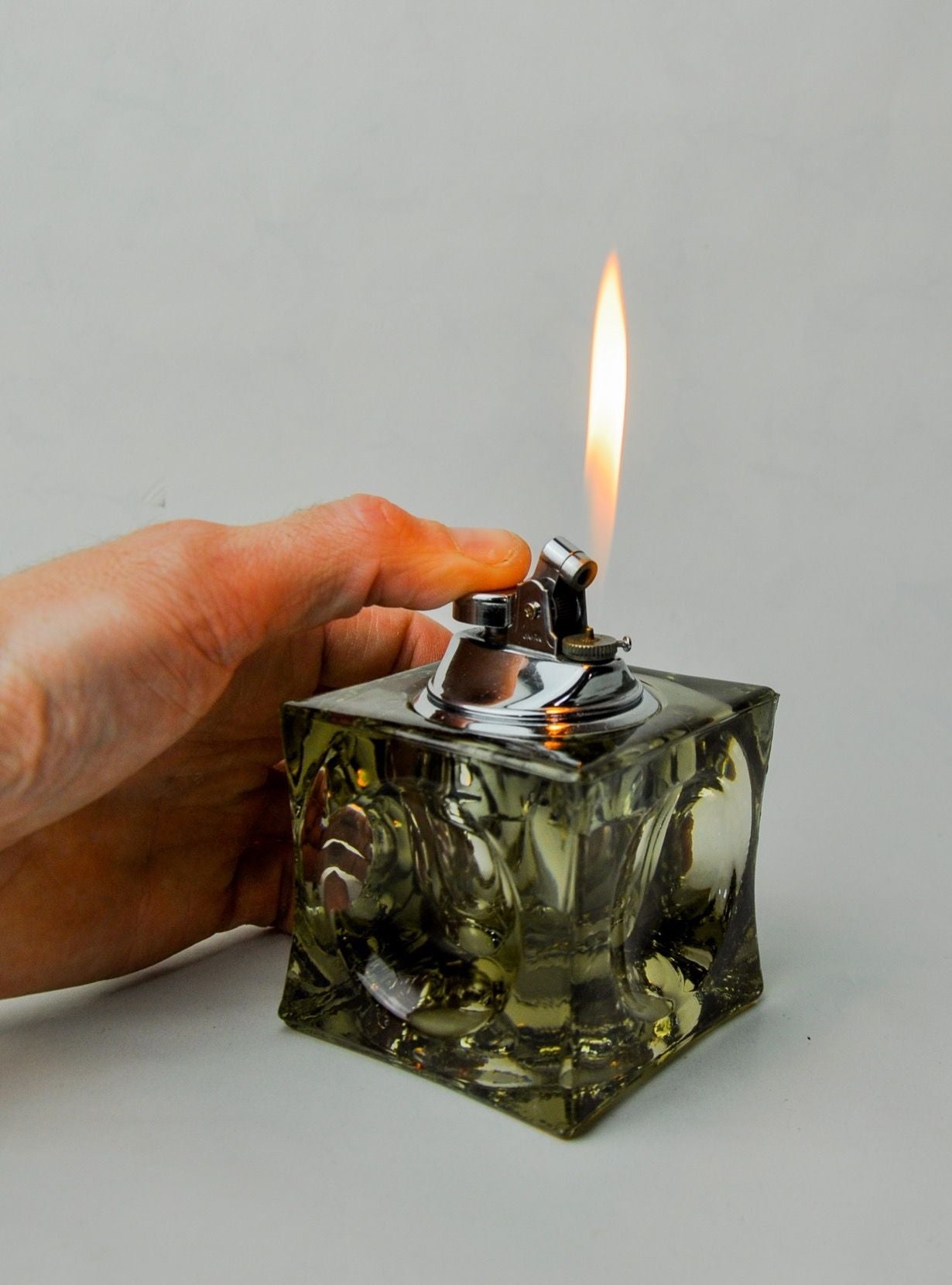 A hand ignites a Malamar Studio ice cube lighter, producing a bright flame from the wick positioned at the top, against a plain light background.