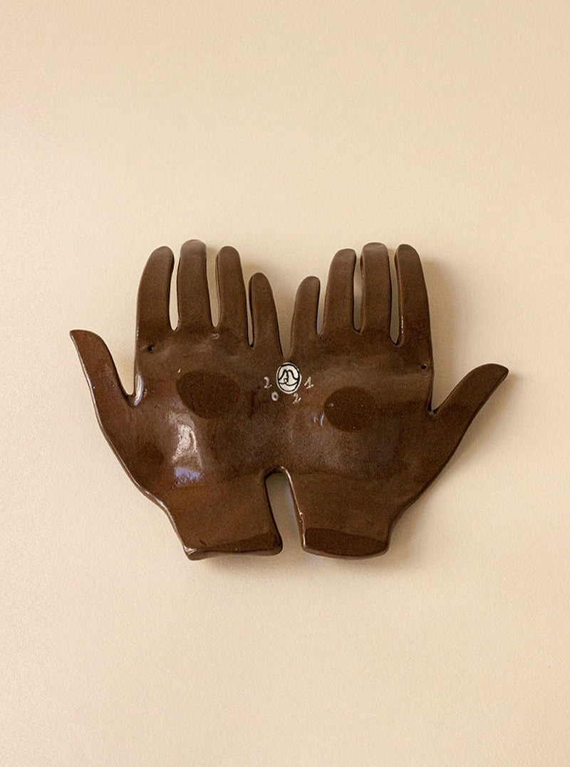 The Main Edition's "The Line" wall piece showcases sculptural ceramic hands with a dark brown glaze, gently cradling the "2021" emblem. These handcrafted hands rest side by side on a light beige background, making it a cherished addition to any collection.