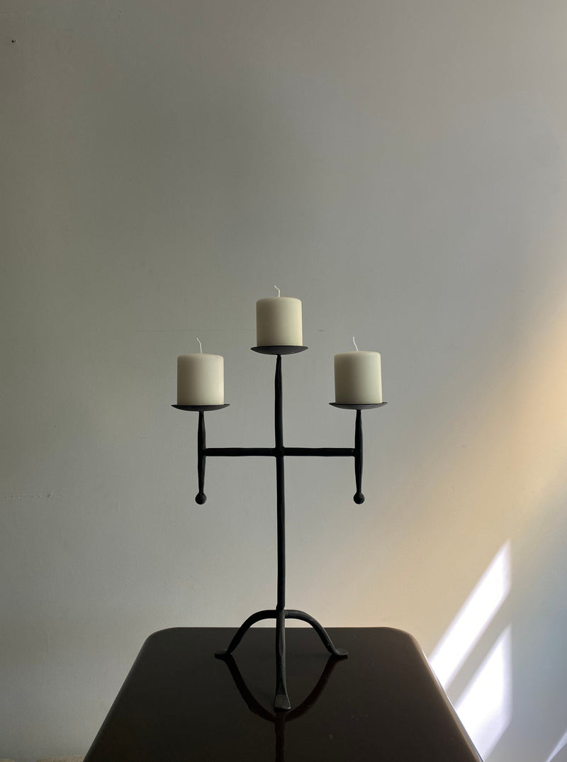 The Vintage Postmodern Iron Candle Holder by Les Objets Amsterdam, with three unlit white pillar candles, stands on a dark table. A soft light casts a shadow on a plain wall behind it, highlighting its contemporary design and creating a serene atmosphere.