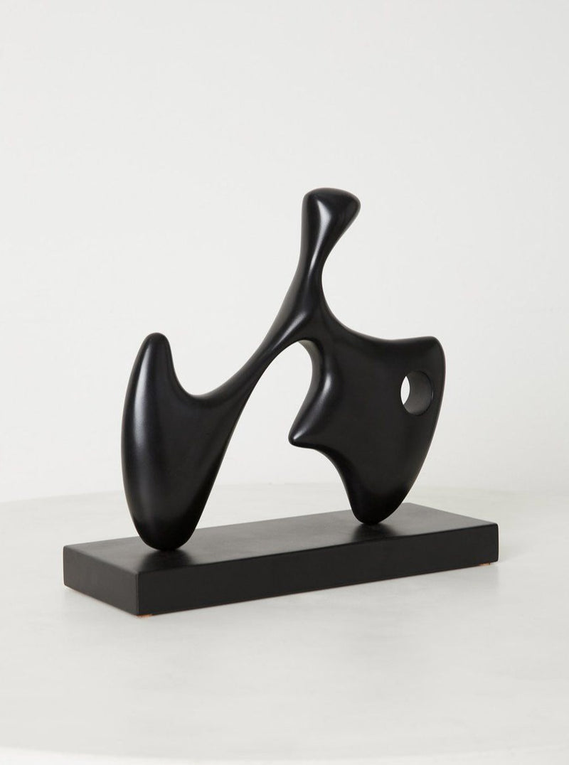 A sleek, black Chandler McLellan Dela sculpture with a fluid, organic shape, featuring curves and a hole, displayed on a rectangular base against a light gray background.