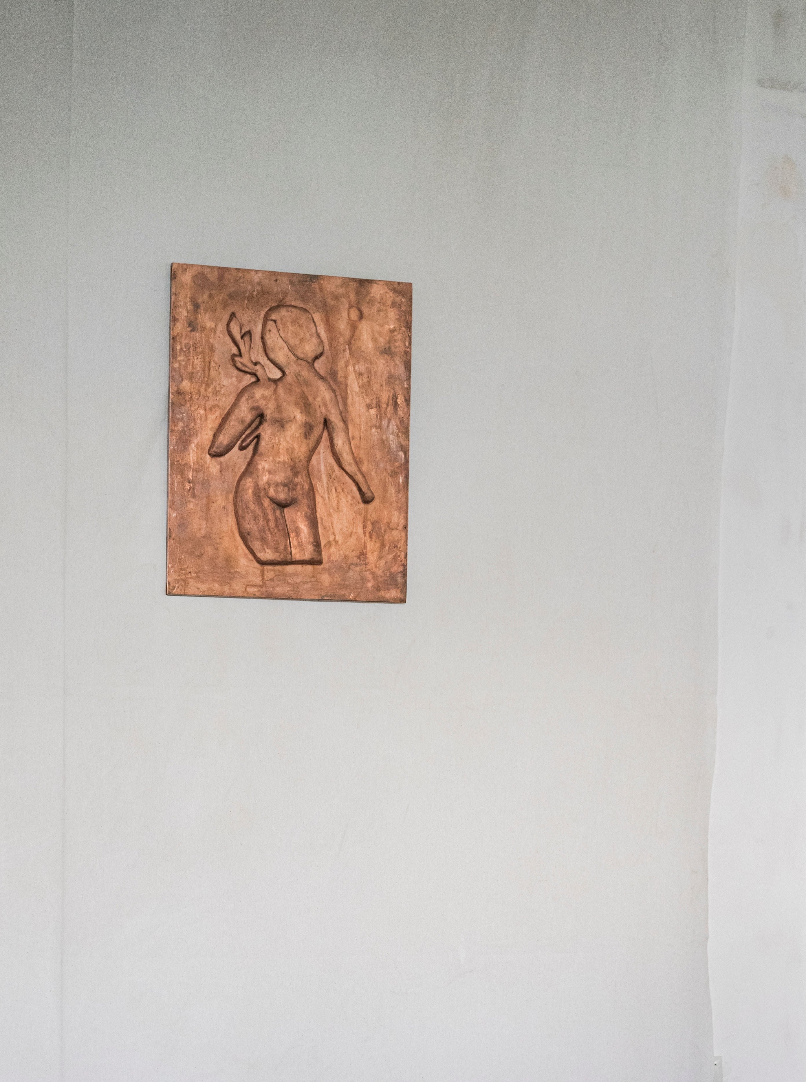 A minimalist artwork of the Woman with Laurel Leaves Chikanka, from Rooms Studio, a stylized female figure with a raised arm, embossed in a copper-toned panel, mounted on a plain, off-white wall with visible imperfections.