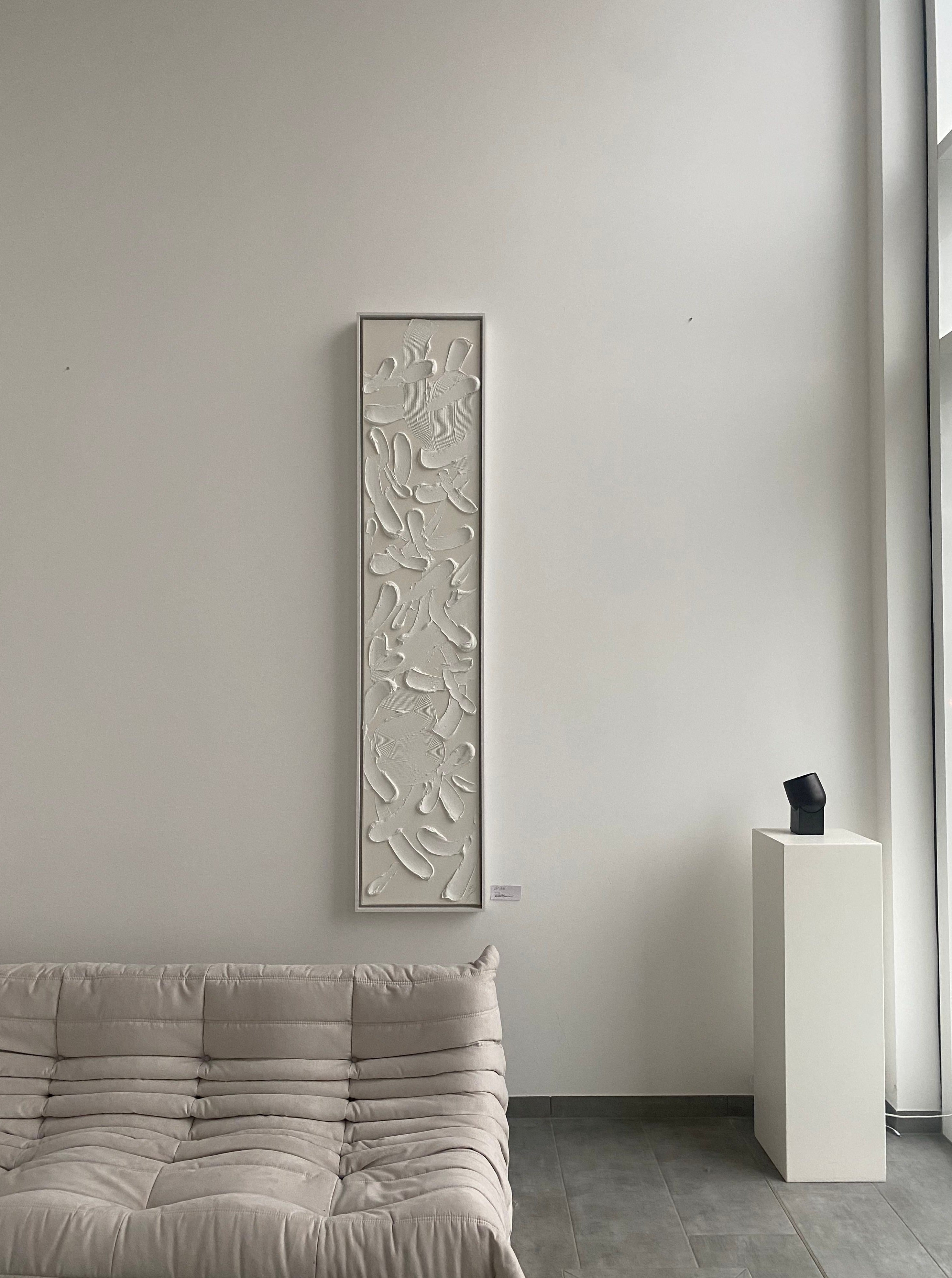 A minimalist room featuring a light gray sofa, a vertically-oriented decorative wall panel with floral motifs, and a black pedestal displaying a "Wild" sculpture by Fulya Celik Art, under natural light.