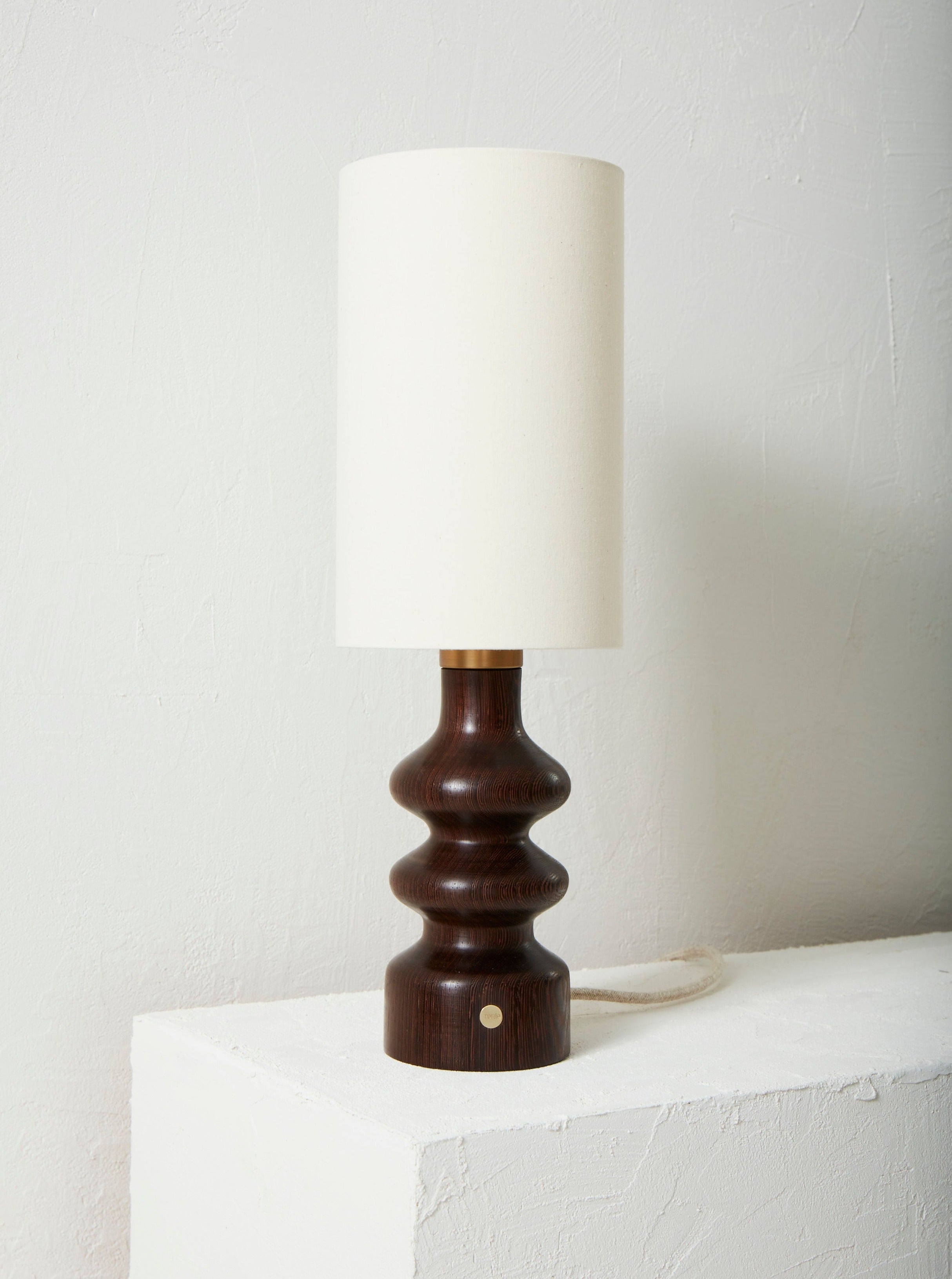 A Alma Wengé Lamp with a textured, wenge grain base and a plain white lampshade, standing on a white shelf against a textured white wall.