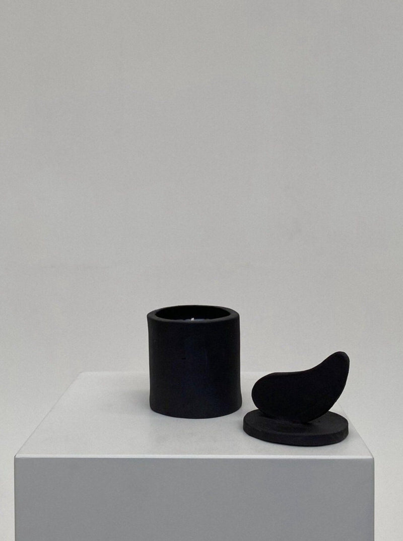 A minimalist display featuring a black cylindrical Sculptural Scented Candle No. 35 holder and a black sculptural object resembling a bird, both placed on a white pedestal against a light gray background.