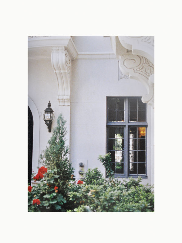 A quaint courtyard entrance with a black door amid white walls, adorned with a hanging lantern and surrounded by lush green plants and bright red flowers, reflects the distinctive Art Deco style of 1920's Maison Plage Preservation of Style.