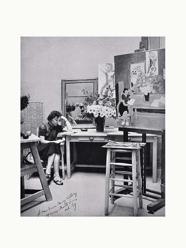 Black and white photograph of a woman seated in an art studio, painting at an easel while surrounded by several canvases and a vase of flowers. A signed inscription by Joan Miro is at theRevu Cahiers d'Art 1940 from Maison Plage.