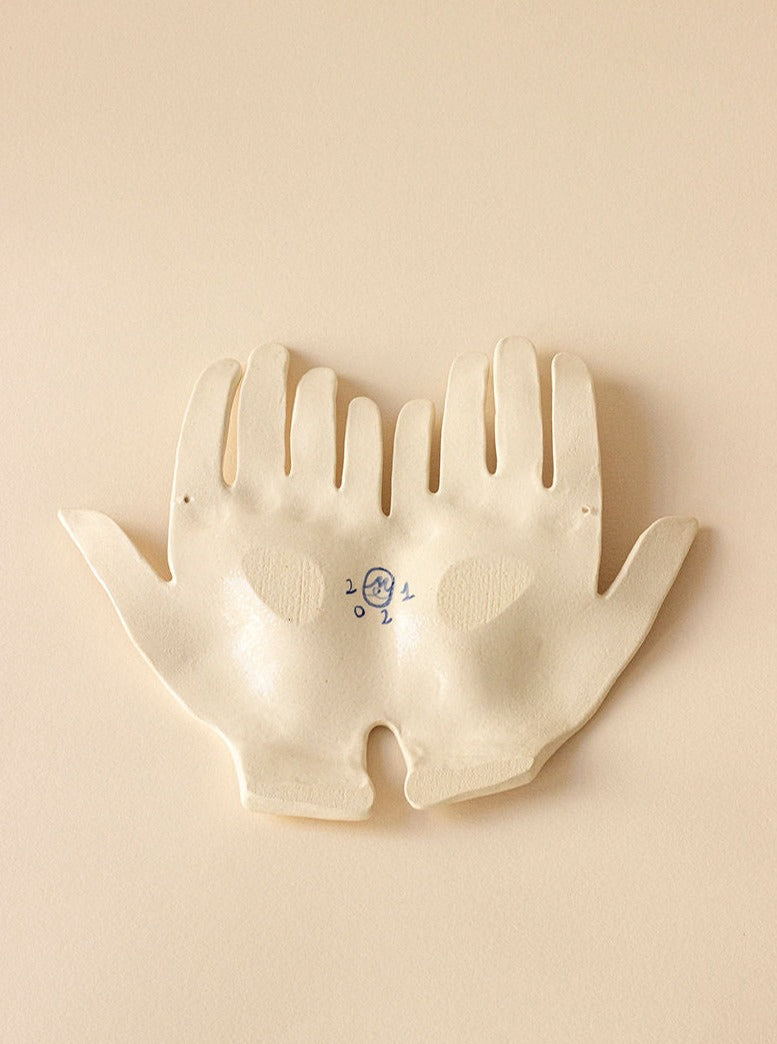The Main Edition's "What binds us" Brown Bond is an exquisite ceramic artwork featuring open hands with heart patterns and small facial features. This off-white piece, resting on a smooth beige surface, embodies contemporary minimalist design for luxury interiors.