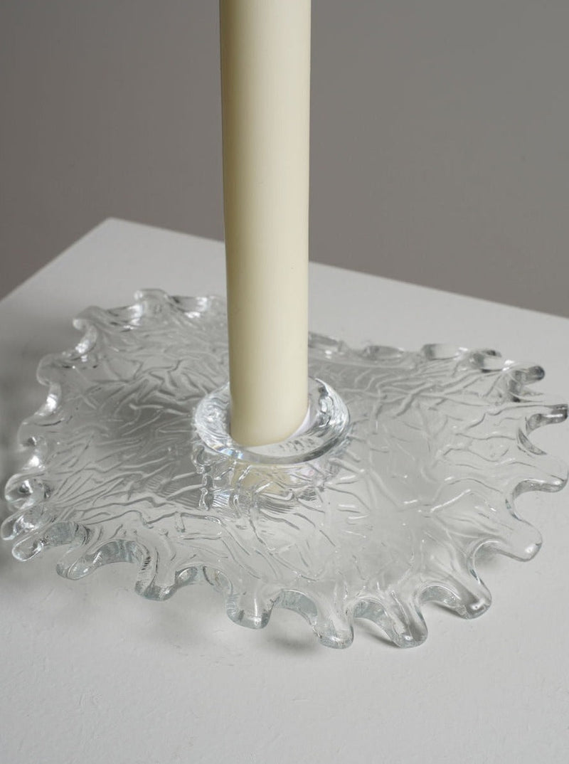 A close-up of a tall, cream-colored candle standing in the intricately designed Uno Westerberg Candle Holder by Les Objets Amsterdam. The glass holder features a wavy, textured edge and sits on a white surface, with a neutral background.