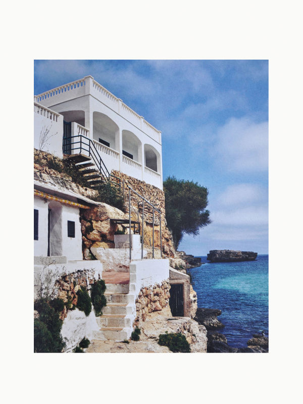 Immerse yourself in the pages of "The Orange Set of 3 Interior Design and Art Books" by Maison Plage, where a white coastal building with stone steps and railings overlooks a tranquil turquoise sea, sparking your creative inspiration. Perched atop a rocky cliff and framed by a partly cloudy blue sky, this picturesque setting also features a nearby rocky outcrop jutting into the water and a solitary tree adding greenery to the serene scene.