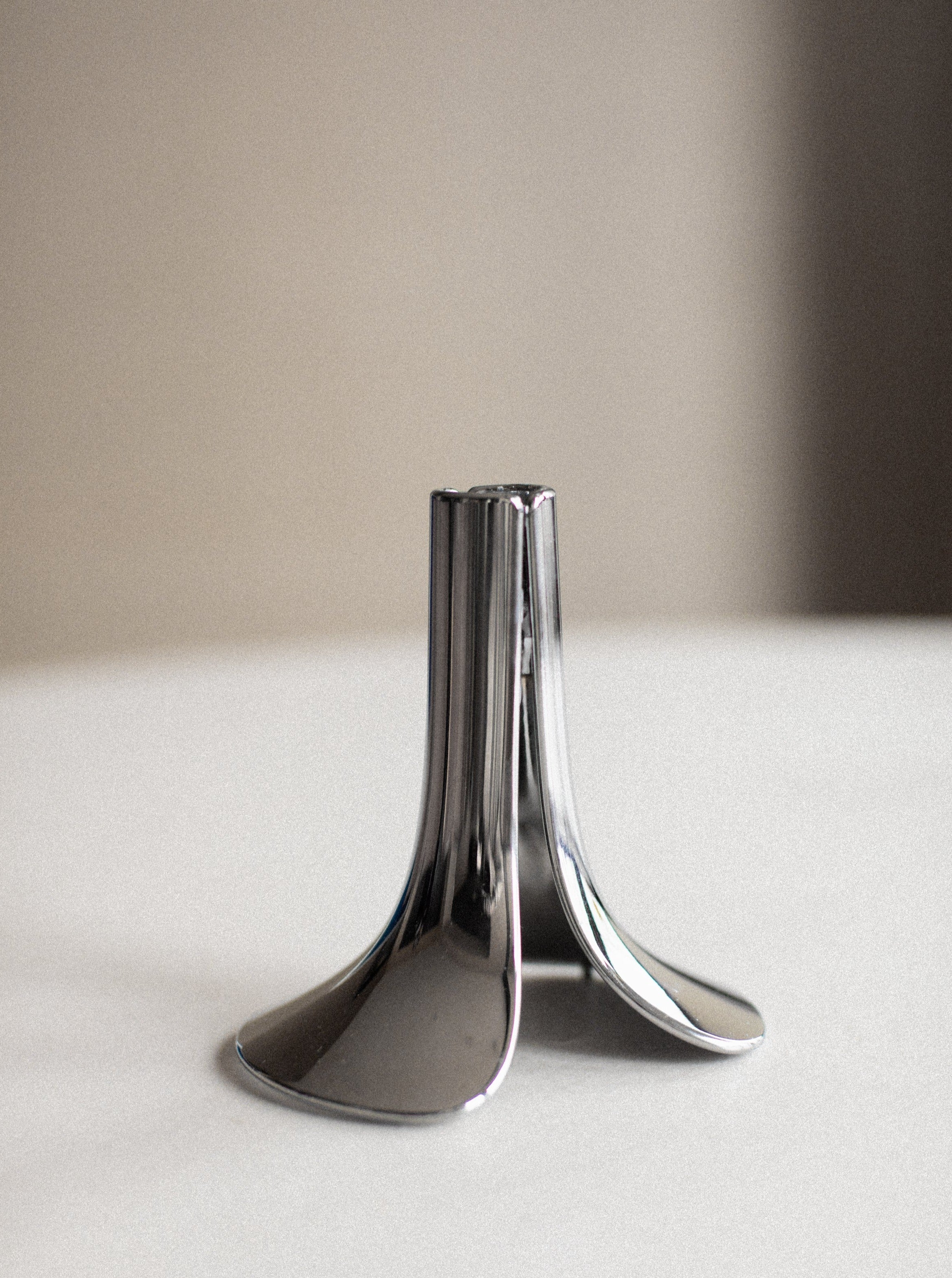 Two shiny Out For Lunch chrome candle holders with a tulip-like design standing on a beige surface against a neutral grey background, highlighting their reflective surfaces and sleek curves.