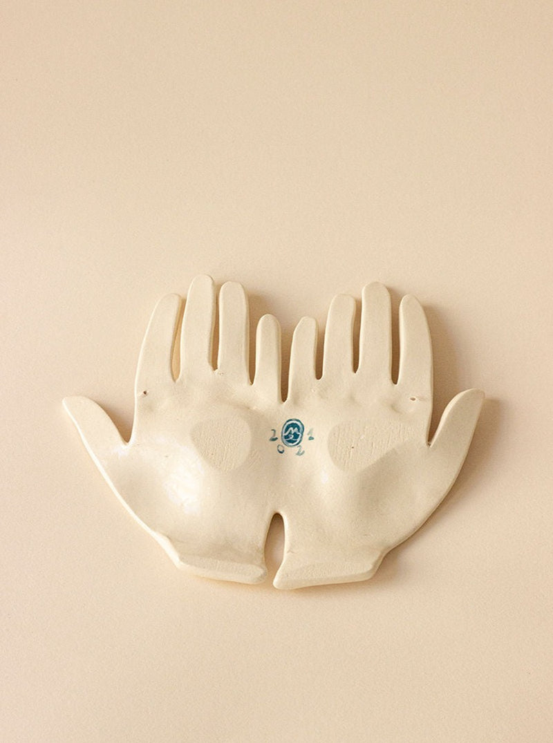 A handmade stoneware dish shaped like two open hands joined side by side, set against a plain, light beige background. The Main Double "What Binds Us" Yellow dish has subtle decorative patterns and a blue emblem on the left palm.