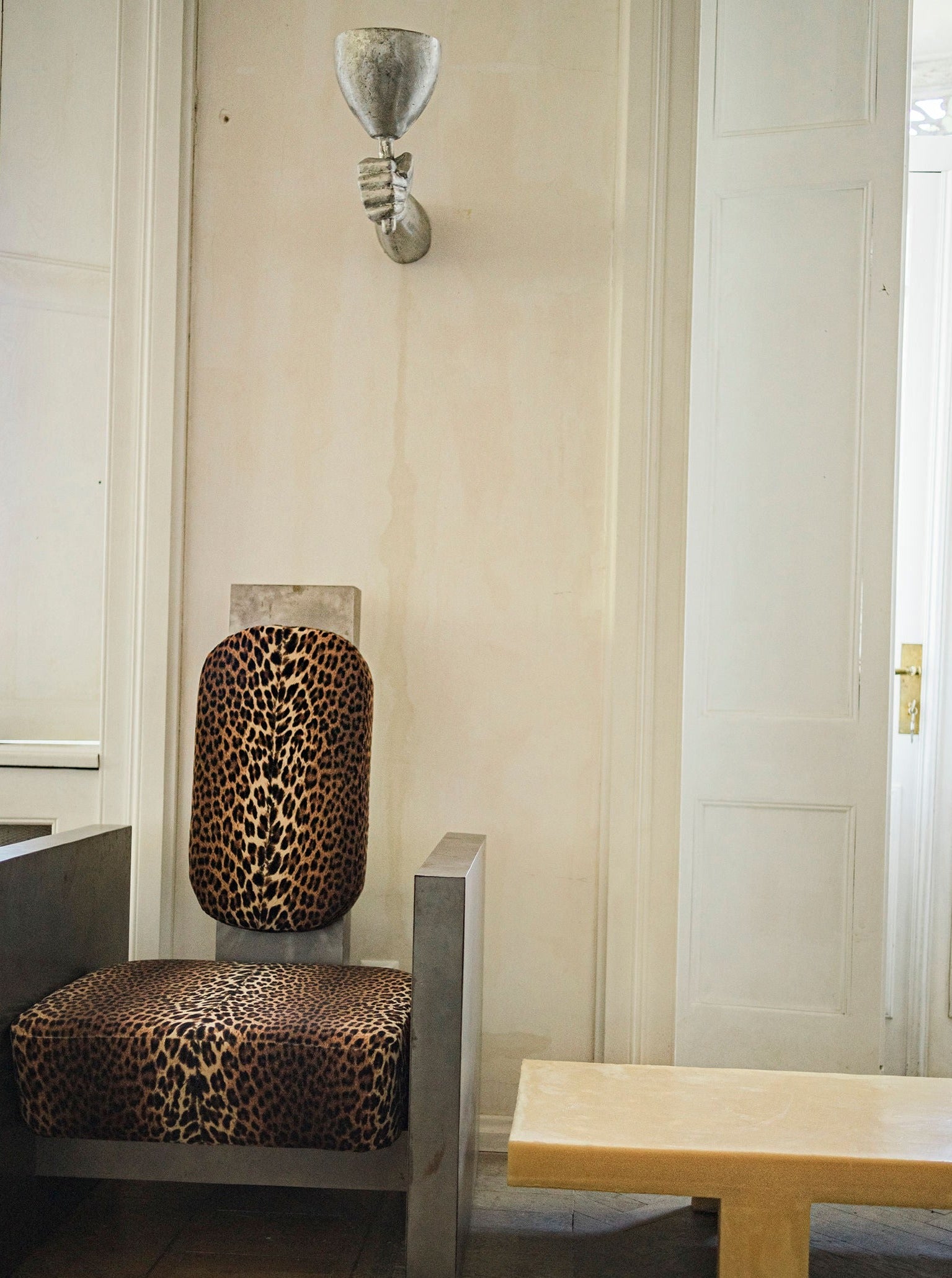 A simple room with minimalist decor featuring an upholstered chair with a leopard print design, a plain wooden bench, and an ornate Rooms Studio Dracula Wall Scone Aluminium.