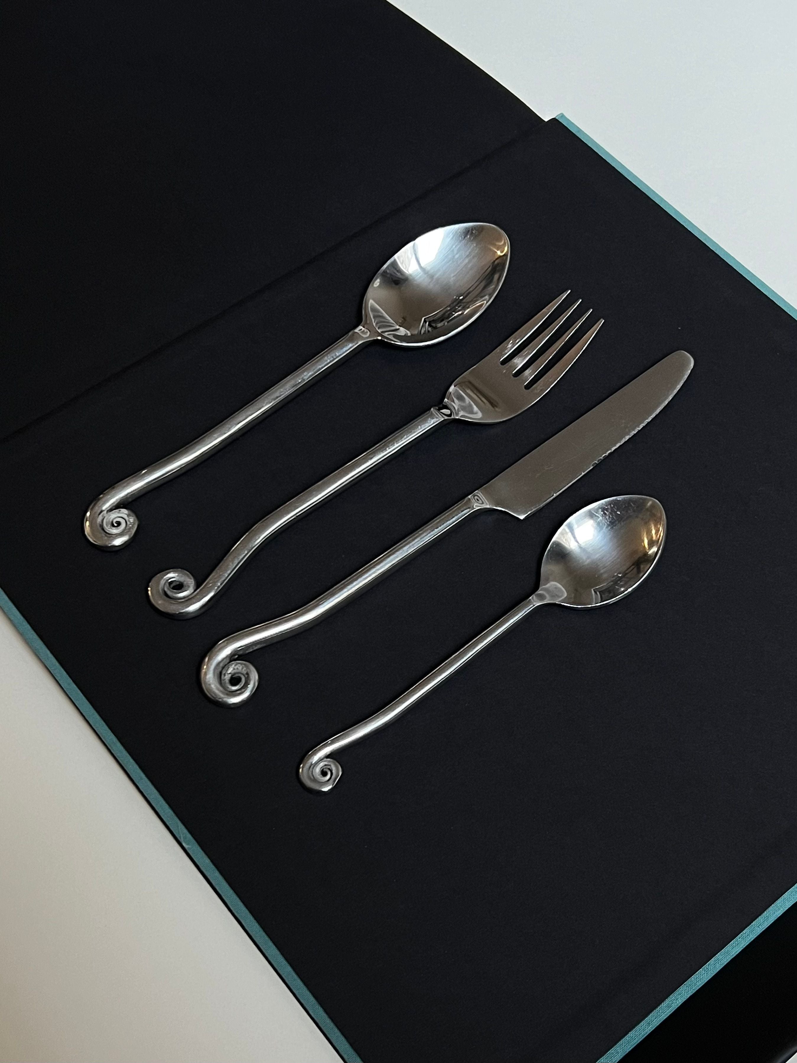 Elegant touch to a Manushi Vintage Swirl Cutlery set consisting of two spoons and a fork with intricate spiral handles, displayed on a dark blue book or box. A minimalist and sophisticated presentation.