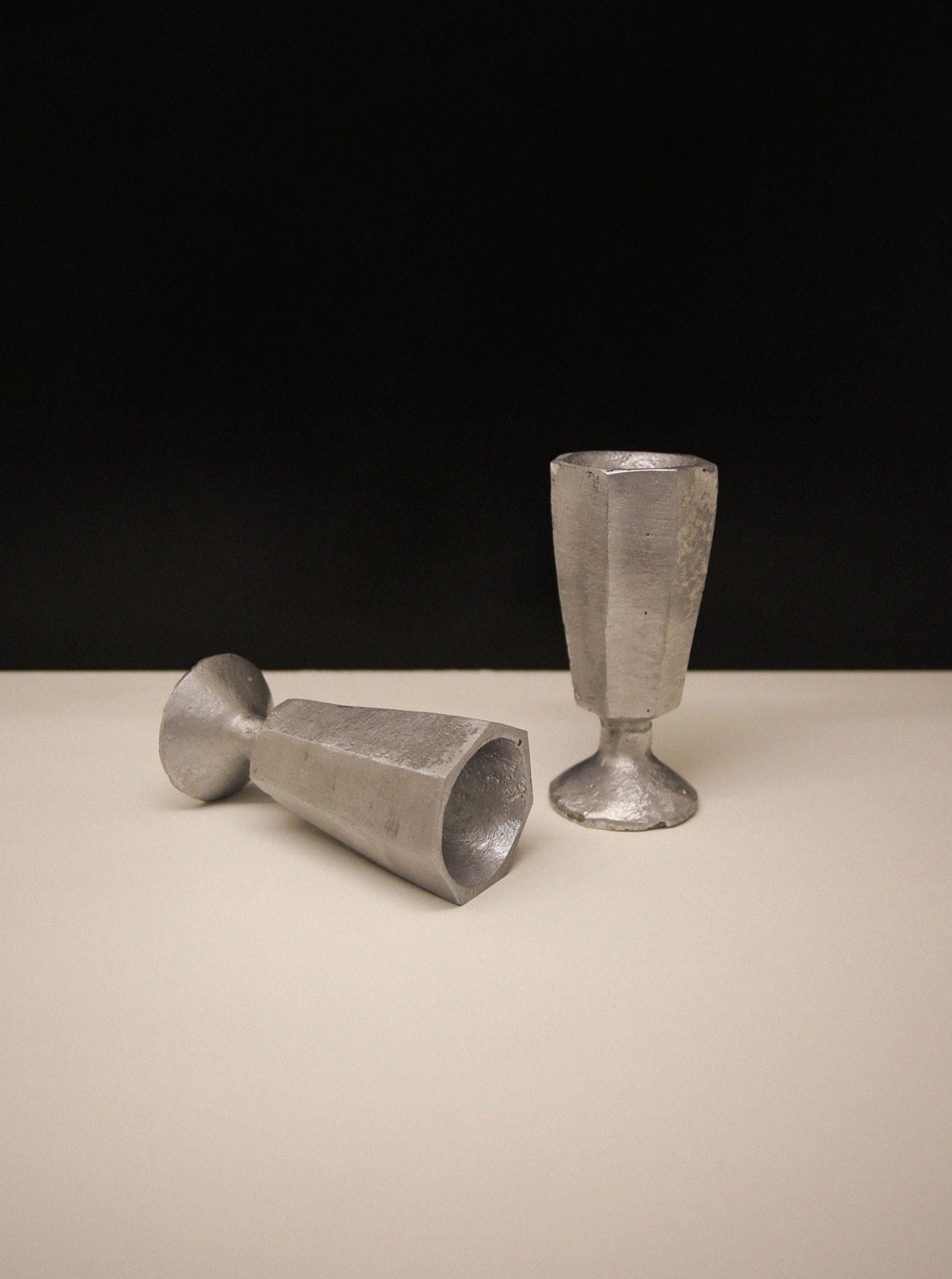 Two Rooms Studio aluminium egg holders, one upright and one lying on its side, on a light surface against a dark background. The egg holders have a rough, handmade texture and serve as elegant egg holders.