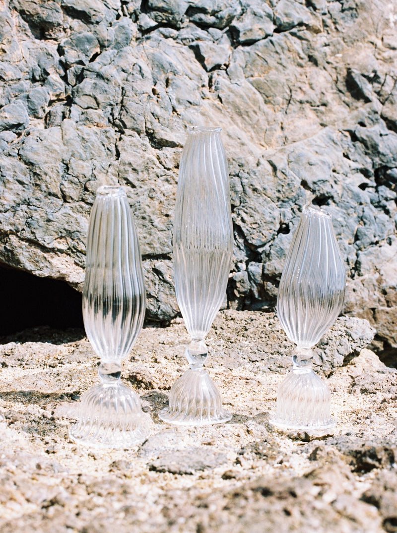 Three The Three Sisters Vases of varying heights standing on a sandy surface against a rugged rock background by Justine Menard.