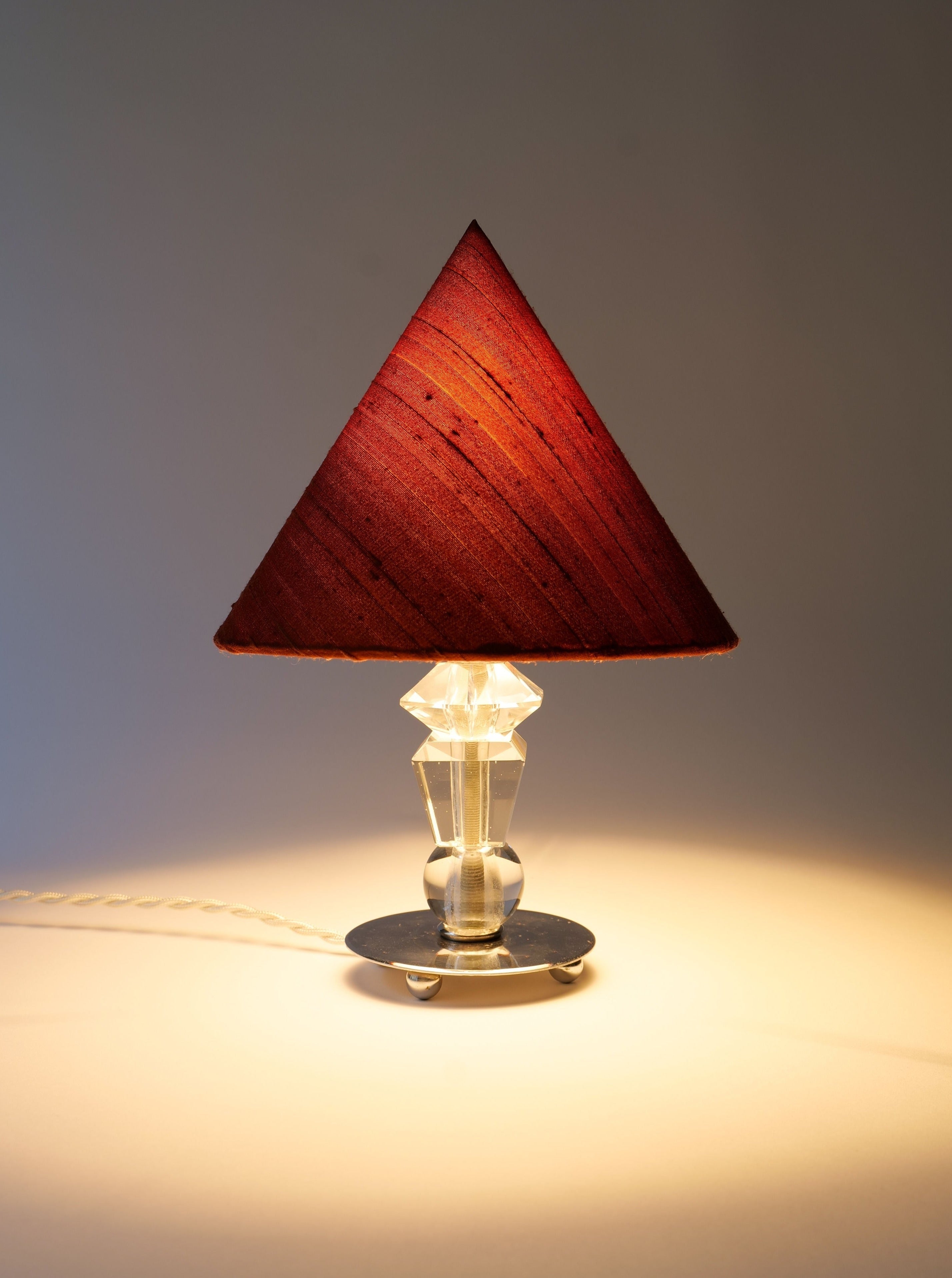A Deco Crystal Table Lamp from the Collection apart with a triangular, red-shaded lampshade illuminated from within, set on a stylized Art Deco silver base, with a lit bulb visible and a subtle glow casting on a plain