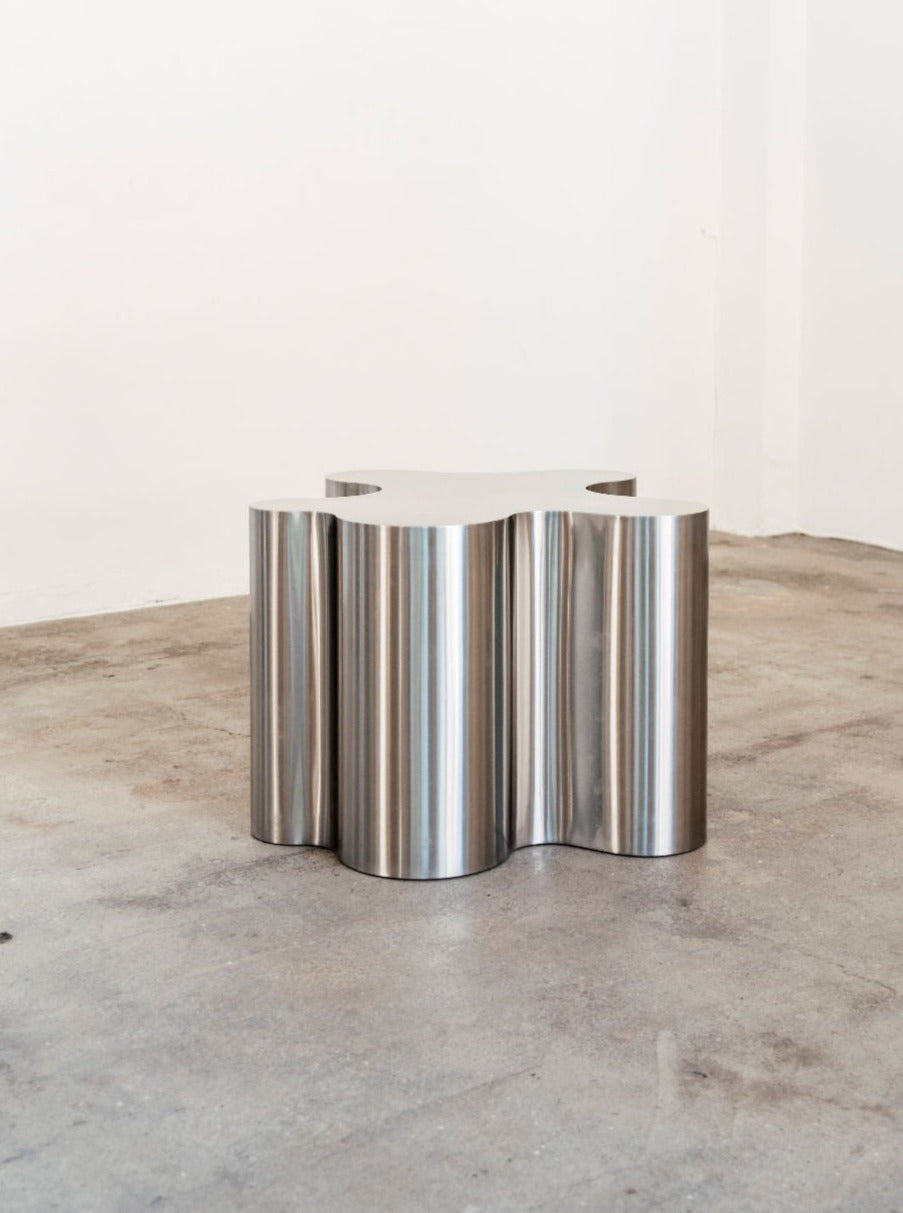 A modern, cylindrical Silver Root - Low side table with a reflective surface and vertical grooves, standing on a concrete floor against a white wall by Caia Leifsdotter.