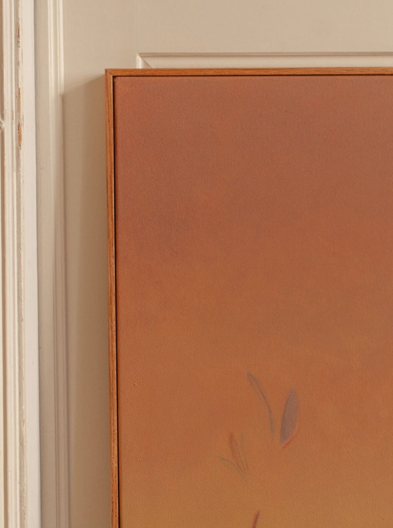 The artwork "Ponds and Breezes #4, 2023" by Rebecca von Matérn features an abstract gradient of warm colors, from brown to dusk orange, with faint leaf-like shapes. It's enclosed in a simple wooden frame with a handcrafted feel, set against the backdrop of a door.
