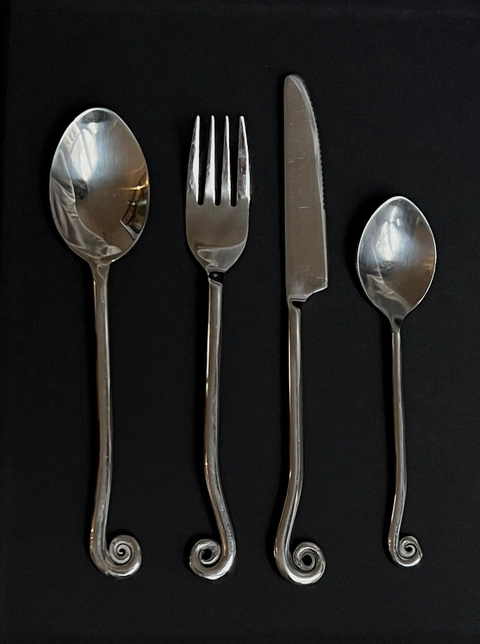 Manushi's Set of Vintage Swirl Cutlery including two spoons, a fork, and a knife with vintage swirl handles, arranged in order on a black background.