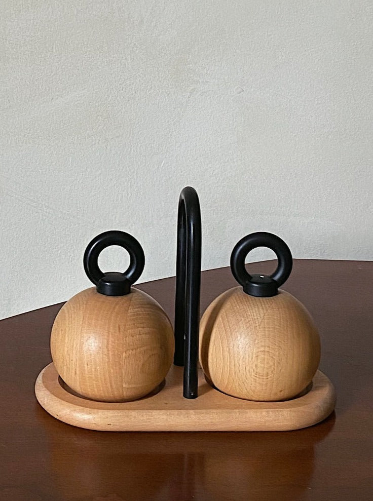 A set of two Bodum C. Jörgensen Salt & Pepper shakers with black handles, designed by C. Jörgensen, placed on a matching wooden tray, against a plain light brown background.