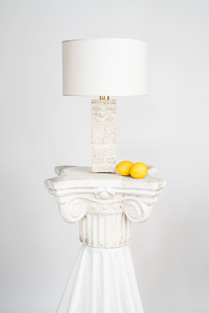 A Capri Travertine table lamp 1 by Brajak Vitberg with a white shade and a unique travertine body sits on an ornate white classical column pedestal, which holds two yellow lemons on a draped white cloth. The background is plain.