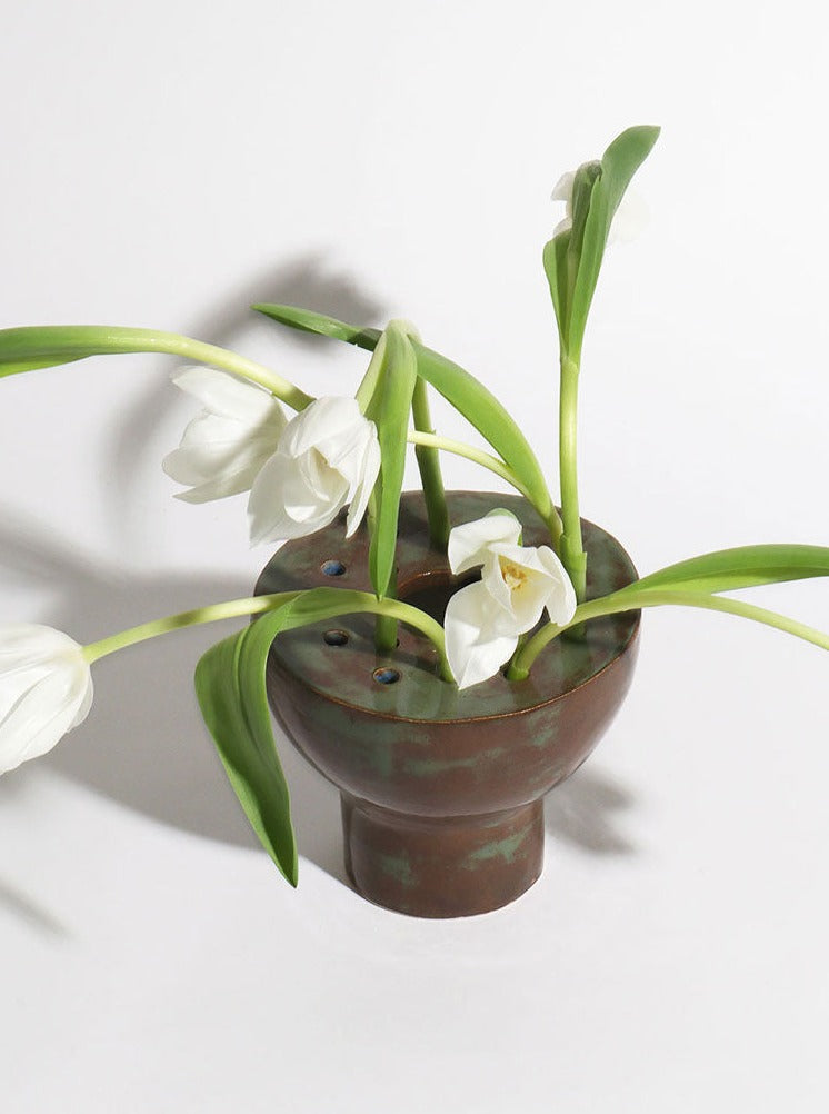 A rustic brown Riola Flora vase with blooming white tulips, gracefully drooping over the edge, against a plain, light grey background.