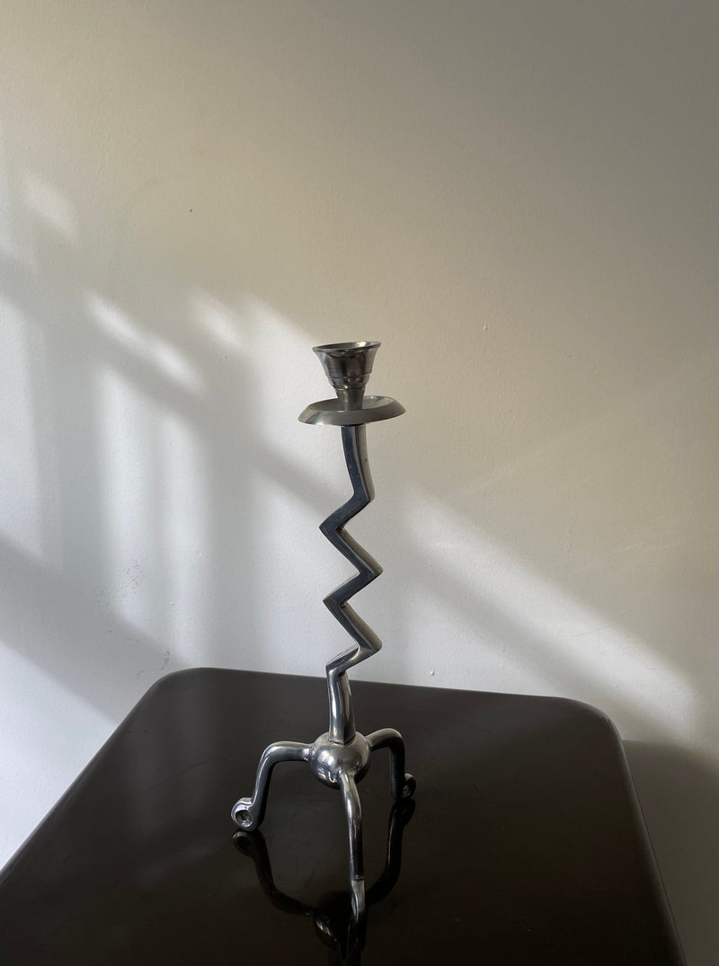 The Vintage Zigzag Candle Holder by Les Objets Amsterdam graces a reflective dark surface against a plain wall, with its zigzag stem and four curved feet. Its sculptural design casts soft shadows in sunlight, adding elegance to luxury interiors.